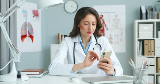 Neşeli gülümseyen kadın doktor portresi. Beyaz önlüklü mutlu genç doktor dolapta oturuyor ve akıllı telefon kullanıyor. Kadın sıhhiyeci çevrimiçi cep telefonuna bağlanıyor, dinleniyor ve geziniyor. — Stok video