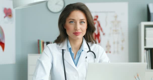 Portrait shot of young beautiful Caucasian woman physician in white gown sitting at table in her cozy cabinet and smiling at camera. Attractive female doctor at laptop computer. Healthcare concept. — Stock Video