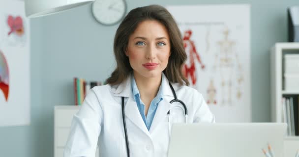 Portrait prise de vue de jeune belle médecin femme caucasienne en robe blanche assise à la table dans son cabinet confortable et souriant à la caméra. Attrayant médecin féminin à l'ordinateur portable. Concept de soins de santé . — Video