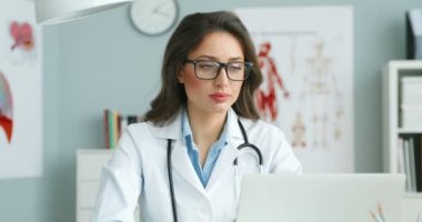 Masasında rahat dolabında oturan, yüzünü kameraya çevirip gülümseyen güzel beyaz kadın hekimin portresi. Laptop bilgisayarında çalışan çekici bir kadın doktor. Sağlık hizmetleri kavramı.