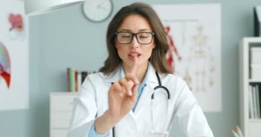 Beyaz önlüklü, gözlüklü, beyaz elbiseli beyaz bir kadın doktor masada oturmuş kameraya sağlık hakkında video blogu yapıyormuş gibi konuşuyor. Kadın sağlık blogcusu sağlık hizmeti ve el sallama hakkında konuşuyor..