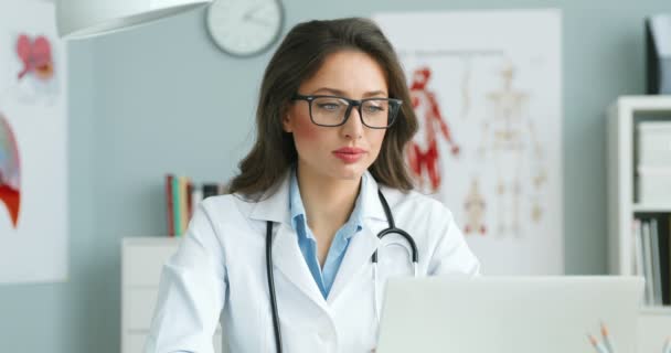 Portret młodej pięknej białej lekarki siedzącej przy stole w przytulnej szafce, odwracającej się twarzą do kamery i uśmiechającej. Atrakcyjna lekarka pracująca przy laptopie. Koncepcja opieki zdrowotnej. — Wideo stockowe