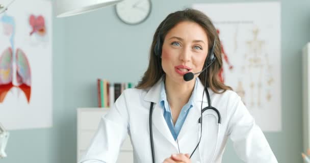 Biała szczęśliwa kobieta lekarz w słuchawkach i białej sukni siedzi przy biurku i rozmawia z kamerą jako wideo o zdrowiu. Radosna kobieta blogerka medyczna mówi o opiece zdrowotnej. Konsultacje z lekarzem — Wideo stockowe