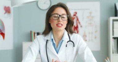 Beyaz önlüklü, gözlüklü, beyaz elbiseli beyaz bir kadın doktor masada oturmuş kameraya sağlık hakkında video blogu yapıyormuş gibi konuşuyor. Kadın sağlık blogcusu sağlık hizmeti hakkında konuşuyor. Hekim danışmanlığı.