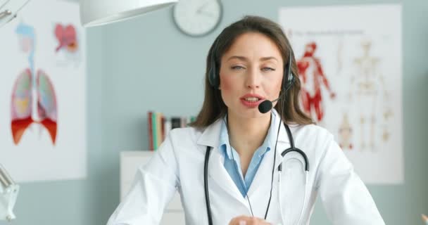 Portret van een blanke jonge vrouwelijke arts in headset en witte jurk zittend aan tafel en pratend met camera als videoblog over gezondheid. Vrouwelijke medische kerel spreekt over gezondheidszorg. Medisch concept — Stockvideo