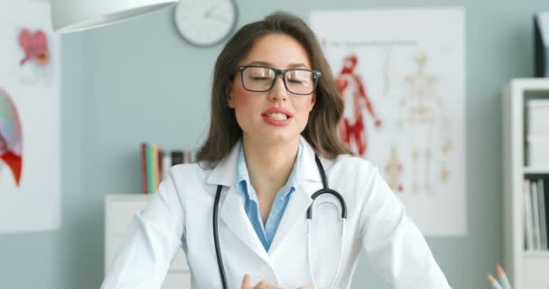 Beyaz önlüklü, gözlüklü, beyaz elbiseli beyaz bir kadın doktor masada oturmuş kameraya sağlık hakkında video blogu yapıyormuş gibi konuşuyor. Kadın sağlık blogcusu sağlık hizmeti hakkında konuşuyor. Hekim danışmanlığı. — Stok video