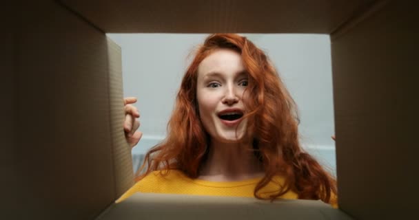 Vista desde el interior de la caja de cartón en la joven mujer blanca alegre con el pelo rojo que mira en ella y sonriendo. Primer plano de bastante feliz chica sonriente apertura caja de cartón. Mujer consiguiendo paquete. — Vídeo de stock