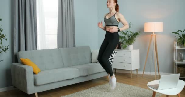 Jonge sportieve vrouw joggen op de plaats in de woonkamer tijdens het doen van fitness thuis tijdens quarantaine. Blank meisje op één plek thuis. Uitwerken bij lockdown zelfisolatie. — Stockvideo