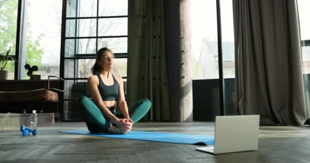 Hermosa blogger deportiva caucásica sentada en la esterilla de yoga en el suelo en casa frente a la computadora portátil. Chica entrenador de fitness grabación videoblog de entrenamiento. Mujer haciendo vlog en estiramiento. Clases en línea . — Vídeos de Stock