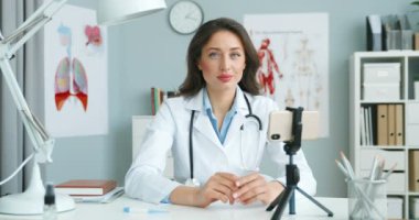 Beyaz cüppeli beyaz kadın doktor portresi dolapta oturuyor ve kameraya bakıyor. Akıllı telefonlu çekici bir kadın doktor. Sağlık blogcusu. Tıbbi günlükler.