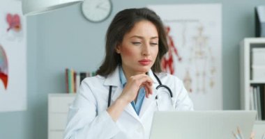 Beyaz elbiseli, beyaz elbiseli beyaz bir kadın doktor dolapta masada oturuyor ve dizüstü bilgisayarda çalışıyor. Güzel kadın doktor ekrana bakıyor ve okuyor. Sağlık hizmetleri kavramı.