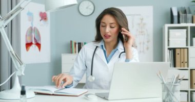 Masasında dizüstü bilgisayarla oturan, cep telefonuyla konuşan ve not defterine randevu yazan bir kadın doktor. Kadın doktor cep telefonuyla konuşuyor ve planlamada danışma zamanına dikkat çekiyor.