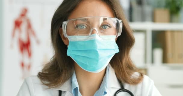 Primo piano del giovane medico caucasico bella donna in maschera medica blu e occhiali, girando faccia a macchina fotografica e guardando dritto Ritratto di bella donna medico indossa protezione da coronavirus — Video Stock