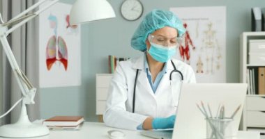 Beyaz önlüklü, tıbbi maskeli, gözlüklü, şapkalı ve eldivenli beyaz kadın doktor dolapta oturuyor ve dizüstü bilgisayar üzerinde çalışıyor. Korunan kadın doktor klavyede yazıyor..