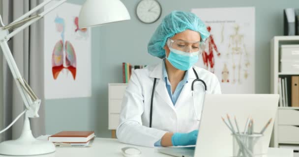 Kaukasische junge Ärztin in weißem Kittel, medizinischer Maske, Brille, Mütze und Handschuhen sitzt am Tisch im Schrank und arbeitet am Laptop. Geschützte Sanitäterin tippt auf Tastatur. — Stockvideo