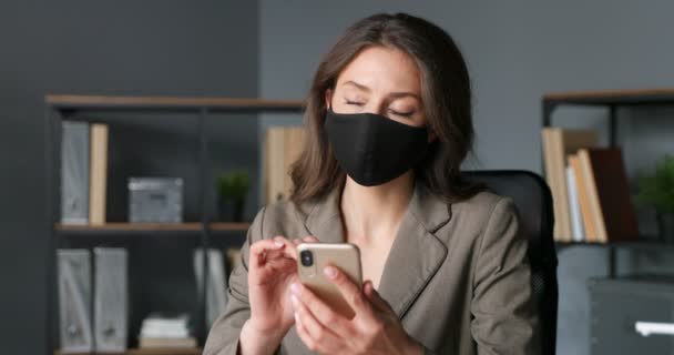 Caucasian young pretty businesswoman sitting in office, texting message on smartphone and dreaming. Beautiful female in mask tapping and scrolling on mobile phone in cabinet. Coronavirus concept. — Stock Video