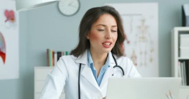Beyaz elbiseli kadın doktor dolapta masada oturuyor ve dizüstü bilgisayarla görüntülü sohbet ediyor. Gülümseyen kadın doktor video blogcusu web kamerasıyla konuşuyor ve sağlık hizmetleri hakkında blog yazıyor. Çevrimiçi danışma.
