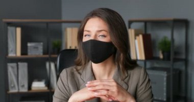 Siyah maskeli güzel beyaz bir kadının portresi ofiste oturuyor ve yüzünü kameraya çeviriyor. İşyerindeki kadın şirket çalışanlarına ya da patronlarına yakın durun. Koronavirüs salgını. İş kadını bakışı.