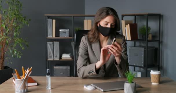 Caucasian young pretty businesswoman sitting at desk in office and texting message on smartphone. Beautiful female in mask tapping and scrolling on mobile phone in cabinet. Coronavirus pandemic time. — Stock Video