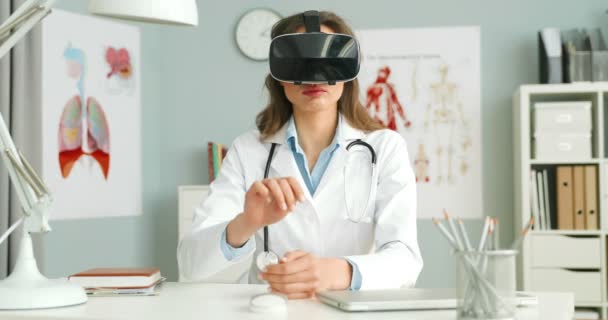 Doctora caucásica en bata blanca y auriculares VR sentada en el escritorio en el gabinete y tocando, desplazándose con las manos en el aire. Concepto de realidad virtual. Salud futurista. Médico trabajando en el futuro . — Vídeos de Stock