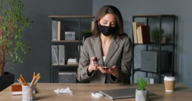 Maskeli şık beyaz kadın masada oturuyor ve ofisteki peçeteyle akıllı telefonu siliyor. Coronavirus salgını koruması. Kadın temizliği, sterilize etme ve cep telefonunu dezenfektanla dezenfekte etme