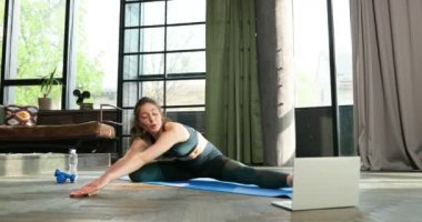 Sportif, beyaz, formda bir kız. Evde, dizüstü bilgisayarın önünde yoga minderinin üzerinde geriniyor. Tecrit çalışması. Online video dersi rehberi. Egzersiz yapan kadın. Blogger rehberi.