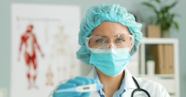 Portrait of young Caucasian woman doctor in white gown, cap, medical mask and goggles showing blurred electronic thermometer to camera. Female medic demonstrating low temperature. — Stock Video