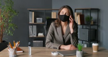 Beyaz, genç, güzel bir iş kadını ofisinde oturuyor ve cep telefonuyla konuşuyor. Maskeli güzel bayan dolapta cep telefonuyla konuşuyor. Coronavirus salgını zamanı. Telefon var..