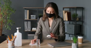 Maskeli güzel beyaz kadın ofis çalışanı masada oturuyor ve dizüstü bilgisayarla fareyi peçeteyle siliyor. Coronavirus pandemik koruma kadın temizliği, sterilize etme ve sterilize etme tekniği