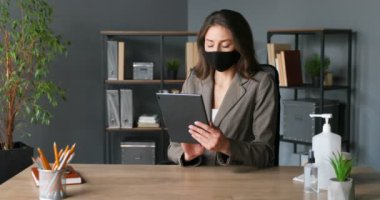 Beyaz tenli genç ve güzel iş kadını ofisinde oturuyor ve tablet cihazını online kullanıyor. Maske takan güzel bir kadın bilgisayar ekranında geziniyor. Koronavirüs salgını.