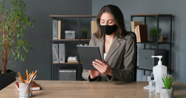 Caucasienne jeune jolie femme d'affaires assise au bureau et en utilisant un appareil tablette en ligne. Belle femelle dans le masque tapant et défilant sur l'écran de l'ordinateur tout en naviguant. Pandémie de coronavirus . — Video