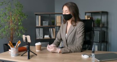 Maskeli şık, beyaz bir kadın ofisinde otururken akıllı telefonunda video kaset taşıyor. Genç iş kadını internet kamerasıyla konuşuyor ve telefonda görüntülü sohbet ediyor. Kadın video iş blogcusu.