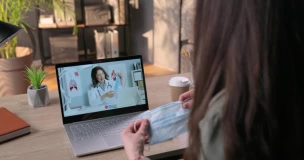 Женщина проводит видеоконсультации с кавказским врачом по поводу медицинской маски. Красивая женщина-врач показывает и объясняет девушке, как использовать маску для лица в онлайн-конференции на ноутбуке. Вирус — стоковое видео
