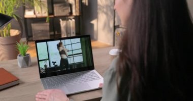 Dizüstü bilgisayarda oturan kız, beyaz kadın fitness antrenörünün kaslarını ne kadar güzel ısıttığını izliyor. Kadın evde karantinada otururken online video çalışması izliyor. Kilitleme kavramı