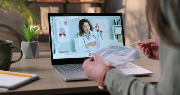 Médico mostrando y explicando a la muchacha, cómo usar la máscara facial en la videoconferencia sobre el portátil. Mujer que tiene consulta en línea con el médico caucásico bastante mujer sobre la máscara médica. Concepto de cuarentena — Vídeo de stock