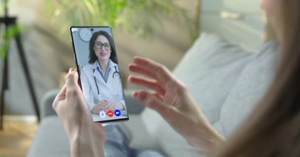 Frau mit Online-Konsultation mit kaukasischen hübschen Ärztin in Video-Chat. Schöne Ärztin spricht mit Mädchen auf Videoanruf auf Smartphone, während sie am Tisch im Krankenhaus sitzt Gesundheitskonzept — Stockvideo