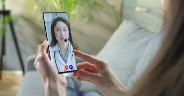 Κορίτσι που έχει ιατρική γνωμάτευση με Καυκάσιο γιατρό σε online βιντεοδιάσκεψη στο κινητό στο σπίτι. Πολύ χαρούμενος γιατρός σε ακουστικά μιλάει με γυναίκα σε βίντεο chat στο τηλέφωνο. Έννοια καραντίνας — Αρχείο Βίντεο