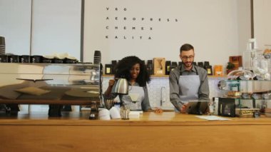 Modern kahve kafede çalışan Afrikalı genç kadın ve beyaz erkek barista, sipariş için dizüstü bilgisayar kullanan müşteri ve adam için kahve yapan kadın..