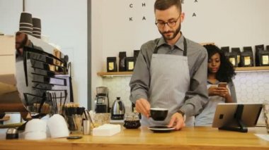 Afrikalı bir kadın akıllı telefondan konuşurken, beyaz tenli genç barista müşteriye taze kahve yapıyor..