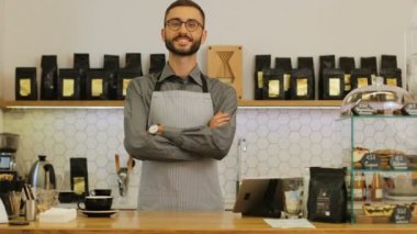 Kahve dükkanında kollarını kavuşturup kameraya gülümseyen baristanın portresi..