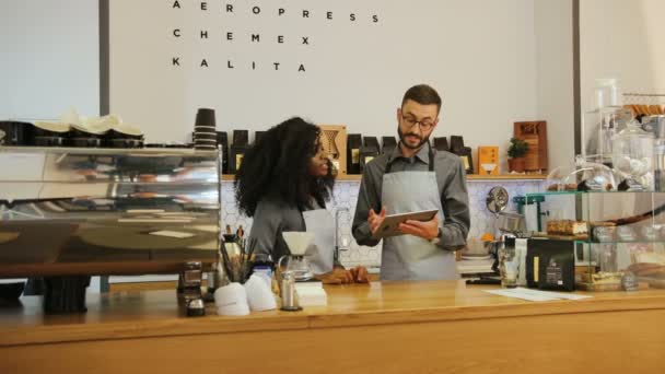 Ung hipster manliga ägare visar afrikansk kvinnlig anställd ny meny av café på tablett under arbetsdagen. — Stockvideo