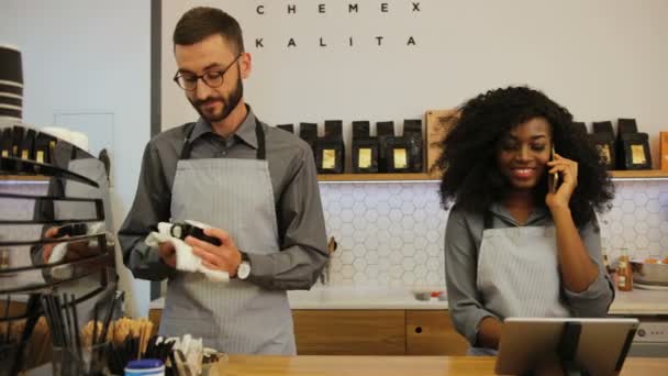 Jonge blanke barista en Afrikaanse jonge vrouw werken in moderne trendy coffeeshop cafe, man wassen beker en vrouw praten op de smartphone. — Stockvideo