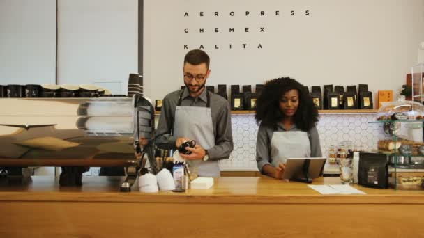 在现代时尚咖啡店的咖啡店、洗碗的男人和使用笔记本电脑的女人中，赫斯特 · 卡斯特（Hipster caucasian）是一位留着胡子、非洲裔年轻女性的咖啡师. — 图库视频影像
