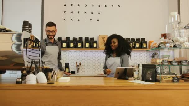 Hipster barista caucásico con barba y mujer joven africana que trabaja en la cafetería moderna de moda. Dos baristas trabajando juntos en el café . — Vídeo de stock