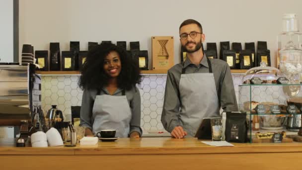 Potret dua barista Kaukasia dan Afrika berdiri di belakang meja kayu ringan, menyilangkan tangan dan tersenyum di depan kamera di kafe. — Stok Video