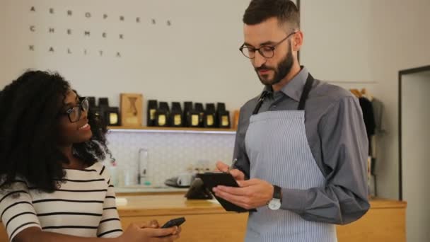 Atractiva mujer africana haciendo un pedido en la cafetería en el camarero hipster, él escuchándola atentamente y anotando todo . — Vídeo de stock