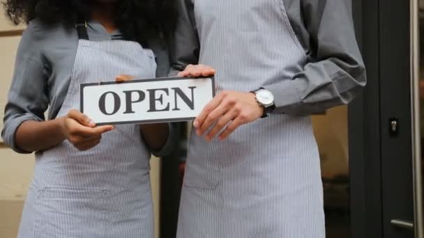 Close up shot of caucasian waiter and african waitress hands holding a board "open" outside the cafe. — Stock Video