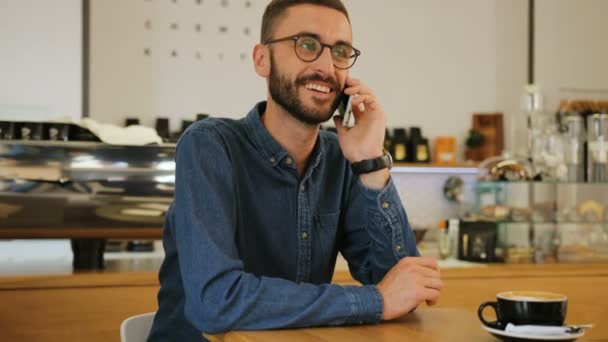 Joven hombre atractivo feliz en los vasos hablando en el teléfono inteligente mientras está sentado en la mesa con una taza de café en la cafetería. Retrato plano . — Vídeo de stock