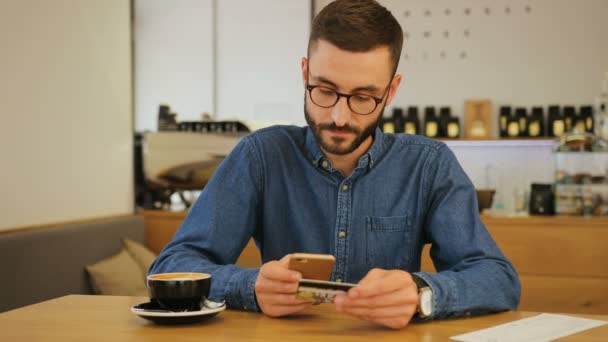 Hipster homem nos óculos usando cartão de crédito para compras on-line pelo telefone inteligente enquanto sittinga em um café . — Vídeo de Stock