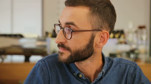 Portrait of young man with beard in the glasses looking to the camera and smiling on camera in the cafe background. — Stock Video
